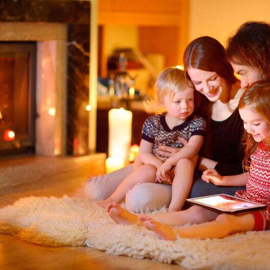 Familie mit zwei Kindern sitzen auf einem Fell umgeben von Kerzen und schauen gemeinsam auf ein Tablet