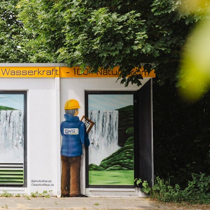 Trafo-Station mit Graffiti: Ein Wasserfall, vor dem ein GWS-Arbeiter mit Helm und Klemmbrett steht, darüber der Text: Die pure Wasserkraft – 100% Naturstrom