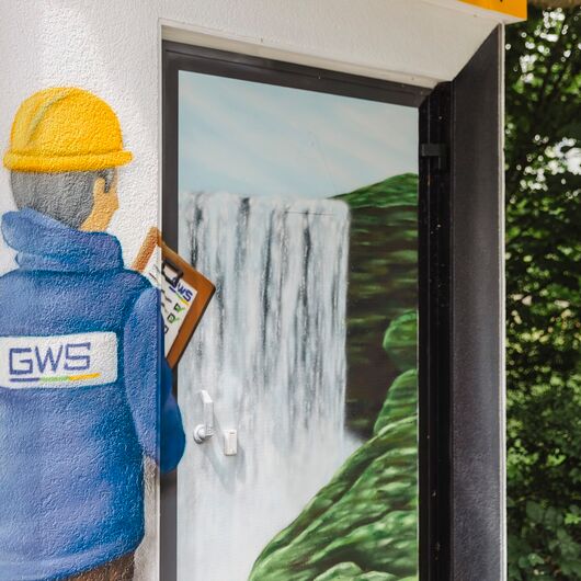 Teil-Aufnahme eines Graffitis, das einen Mitarbeiter mit GWS-Jacke, Helm und Klemmbrett zeigt, der auf einen Wasserfall schaut
