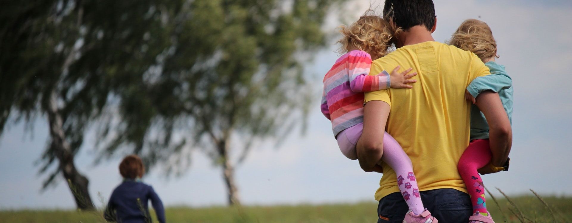 Mann von hinten mit zwei Kindern auf dem Arm, der über eine Wiese läuft, weiter vorne rennt ein drittes Kind